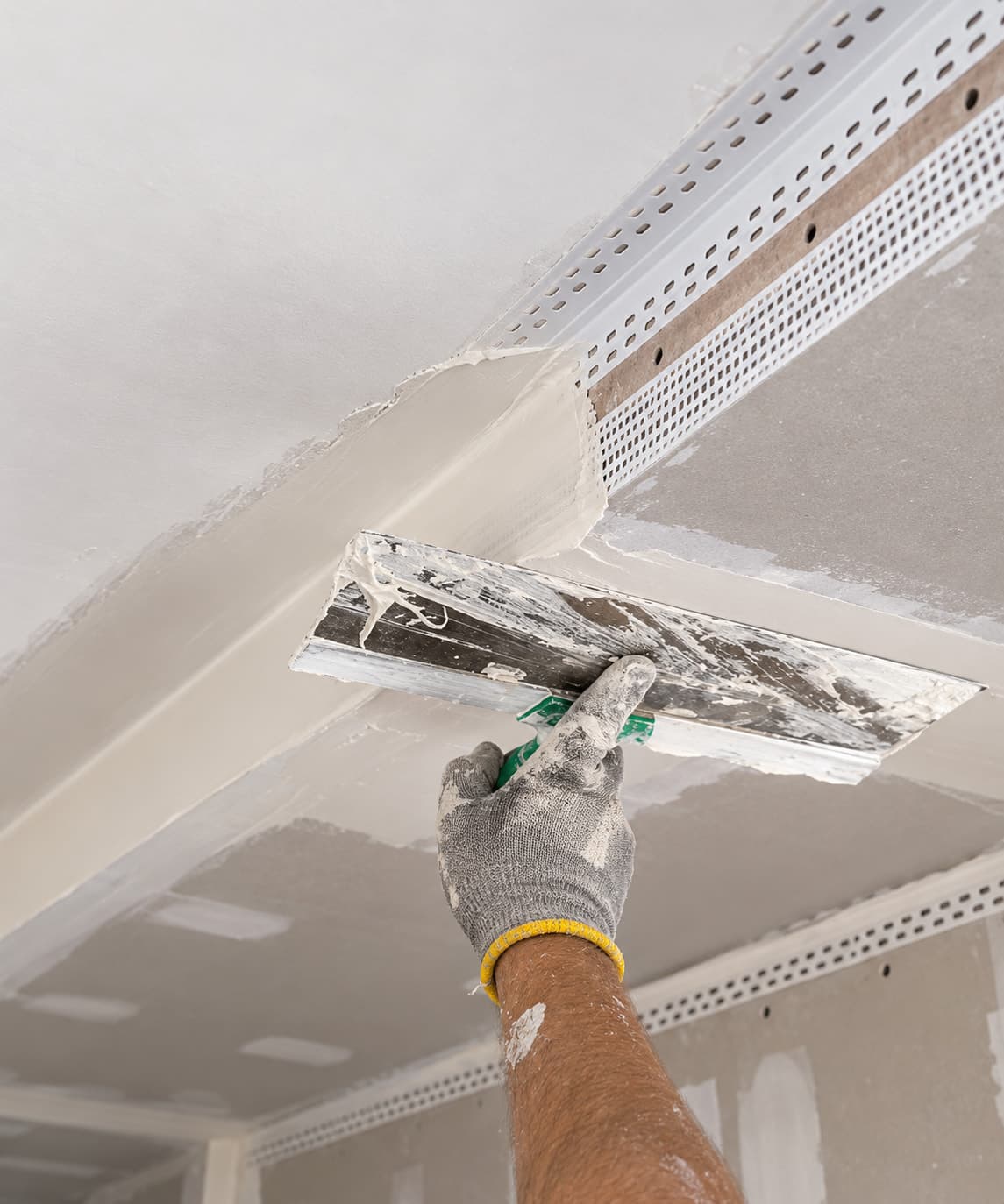 Gloved hand using a wide taping knife to apply joint compound over a perforated bead at a ceiling transition