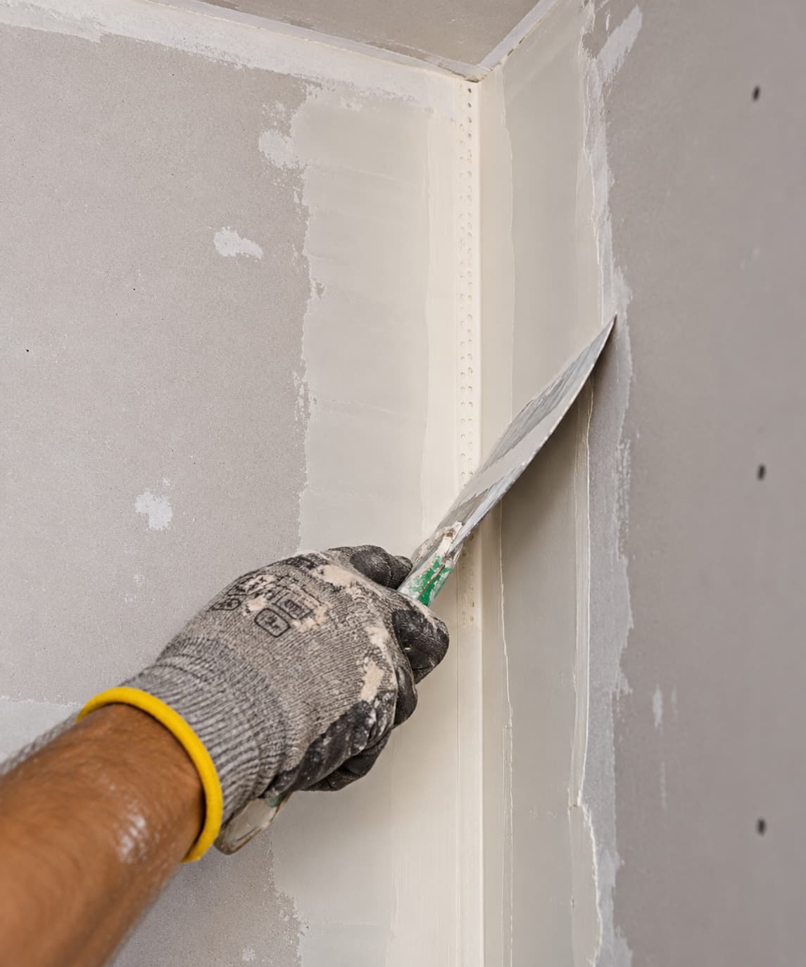 Gloved hand using a putty knife to apply joint compound along an inside corner with a corner bead